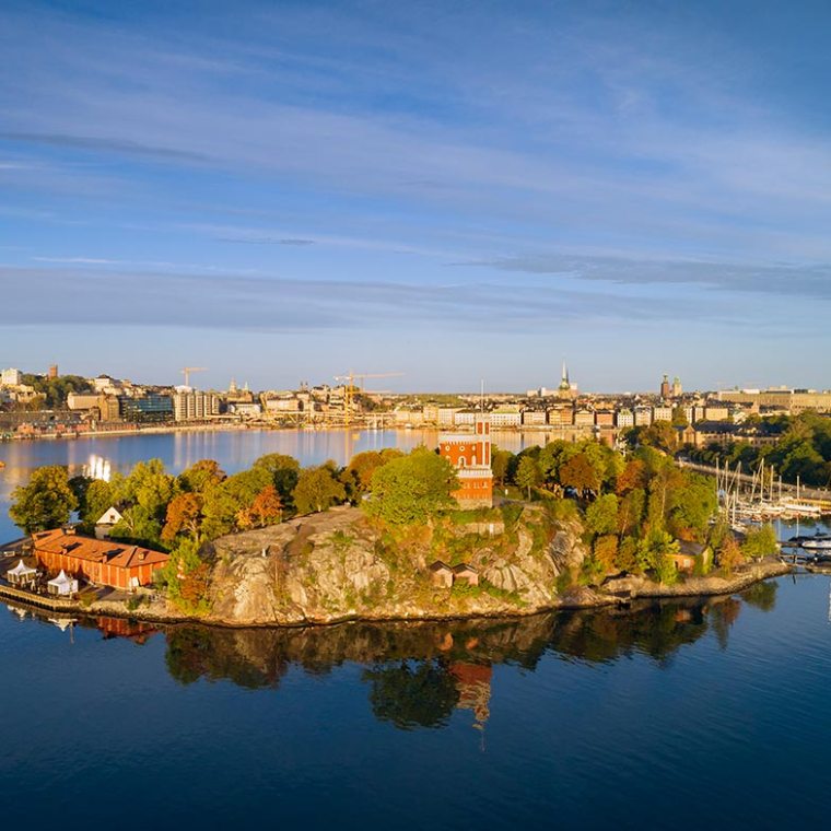 Scenic view of small island with trees and buildings, city skyline in background, calm water reflections.
