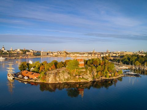 Scenic view of a small island with colorful trees, a red building, and sailboats, surrounded by calm water.