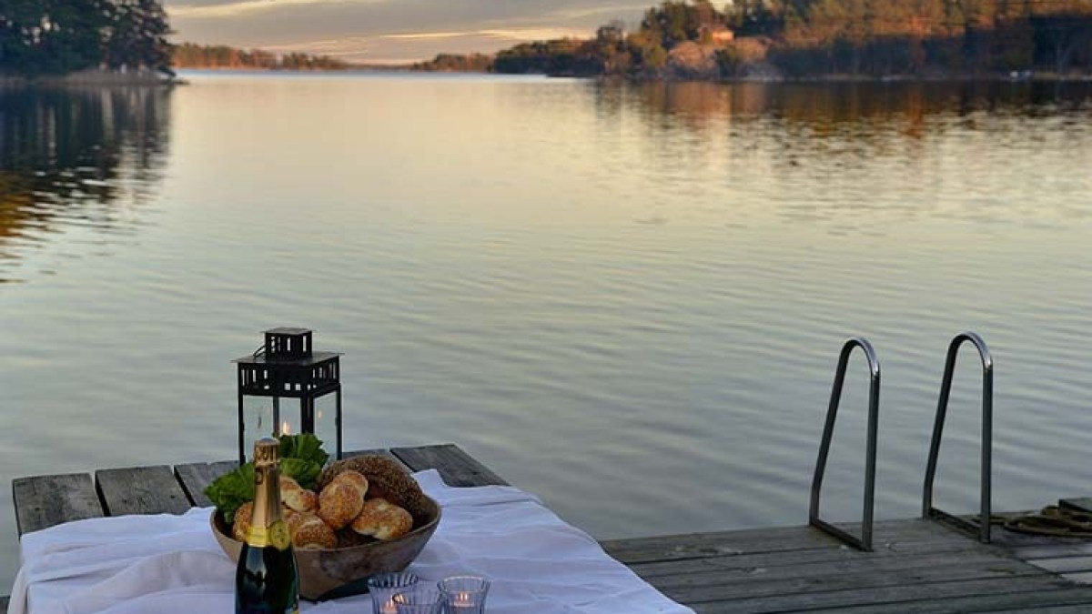 Table with bread, champagne, and candles on a dock by a tranquil lake at sunset.