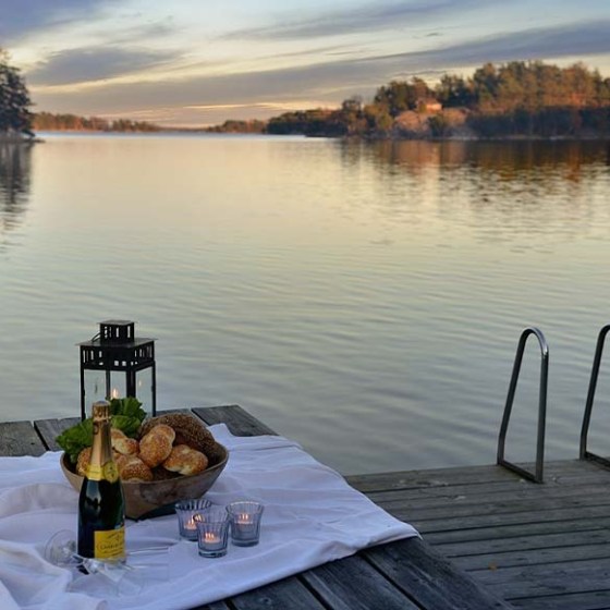 Table with bread, champagne, and candles on a dock by a tranquil lake at sunset.