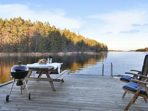 Lakeside deck with grill, picnic table, and chairs overlooking calm water and forest.