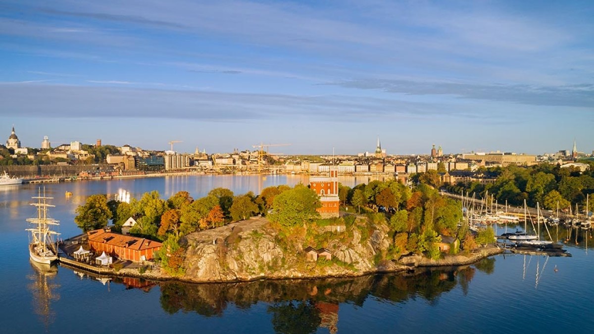 Aerial view of a small island with trees and buildings in a harbor, with a city skyline in the background.