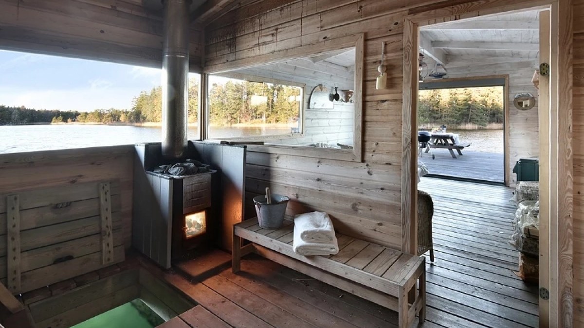 Wooden sauna interior with a stove, lake view, and bench with towels.