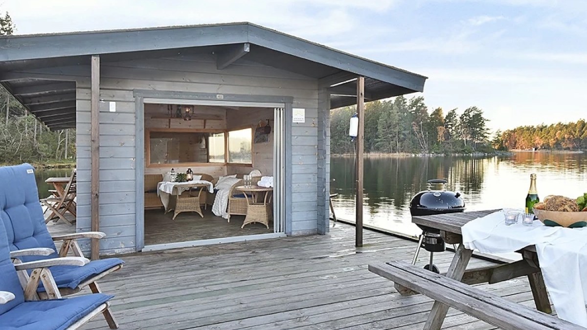 Lakeside cabin with open door, dining table inside, outdoor seating, grill, and picnic table on wooden deck.