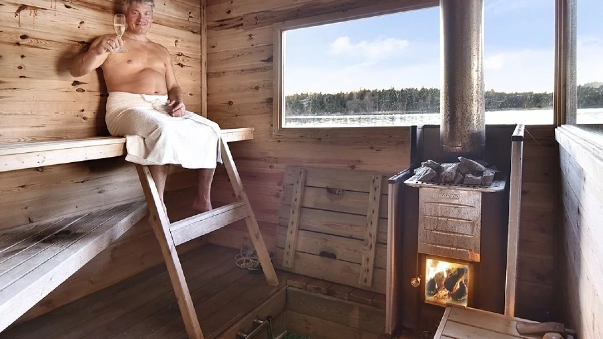 Person seated in a wooden sauna, wearing a towel and holding a drink, with a view of a lake outside the window.