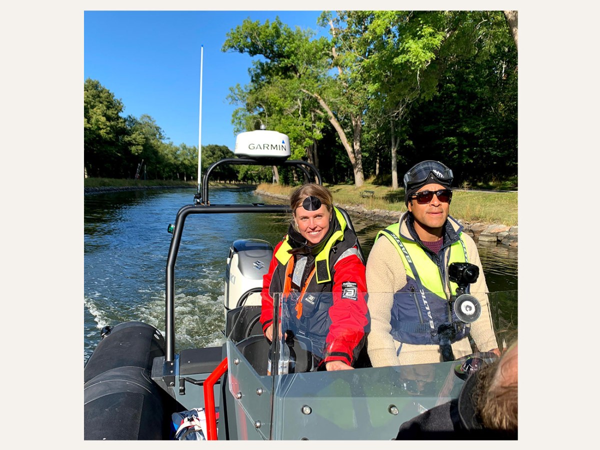Two people in life jackets driving a small boat on a sunny waterway.