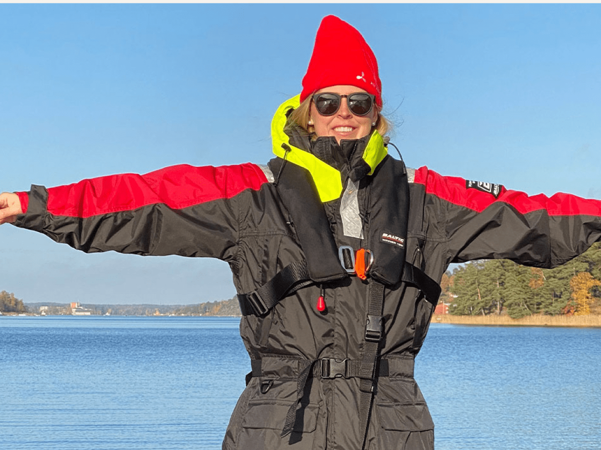 Person in red and black jacket with outstretched arms, wearing a red hat and sunglasses by a lake.