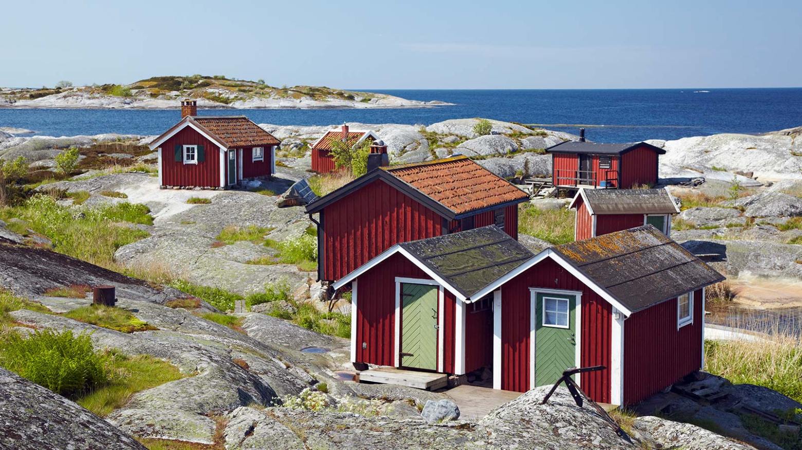 Red cabins on rocky shore with sea and islands in background under a clear sky.