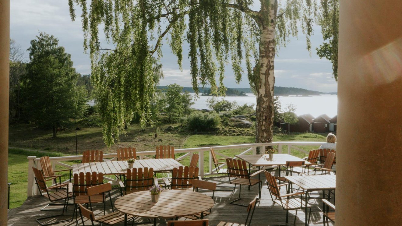 Outdoor patio with wooden tables and chairs under a tree, overlooking a lake and greenery.