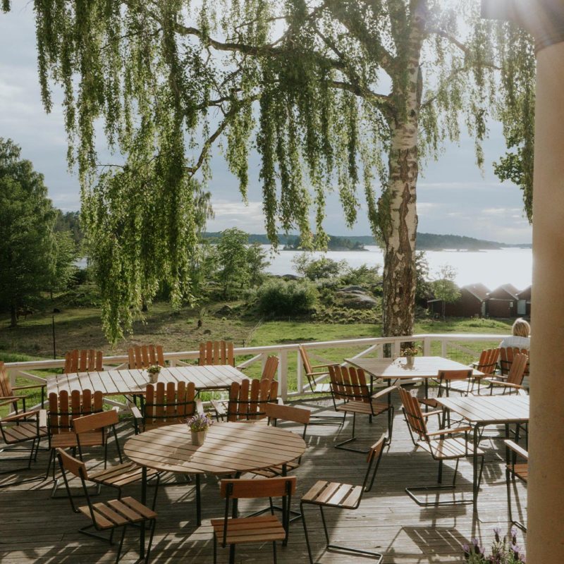 Outdoor patio with wooden tables and chairs under a tree, overlooking a lake and greenery.