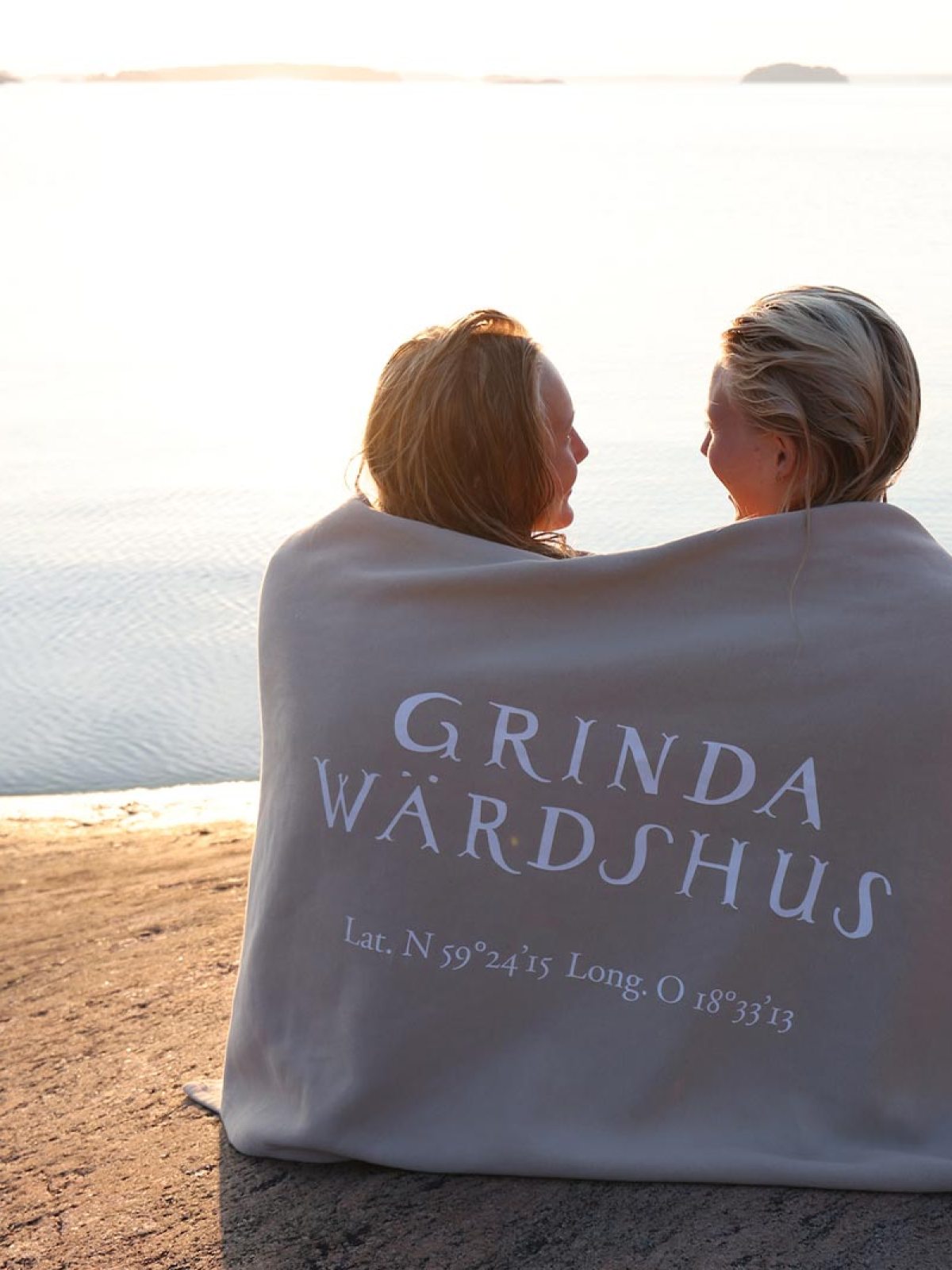 Two people wrapped in a blanket sitting by a calm lake at sunset.