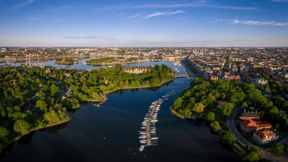 Aerial view of a cityscape with green parks, water, boats, and buildings under a clear blue sky.